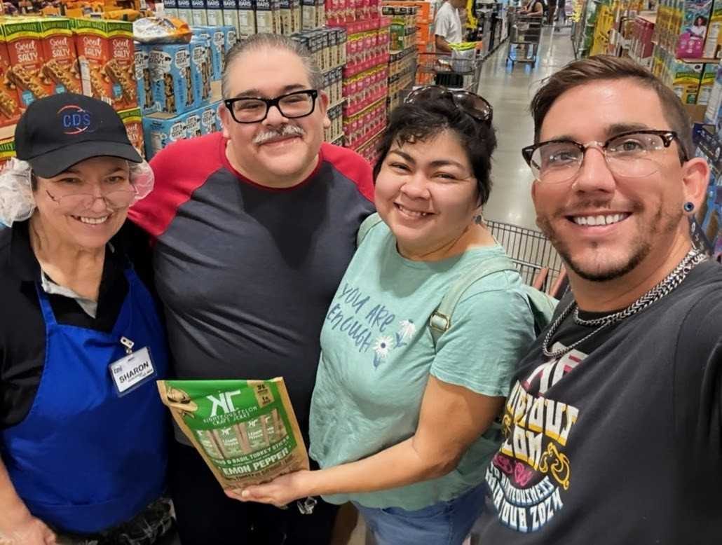 Four people in a grocery aisle standing shoulder to shoulder, holding a product box up to the camera and smiling.