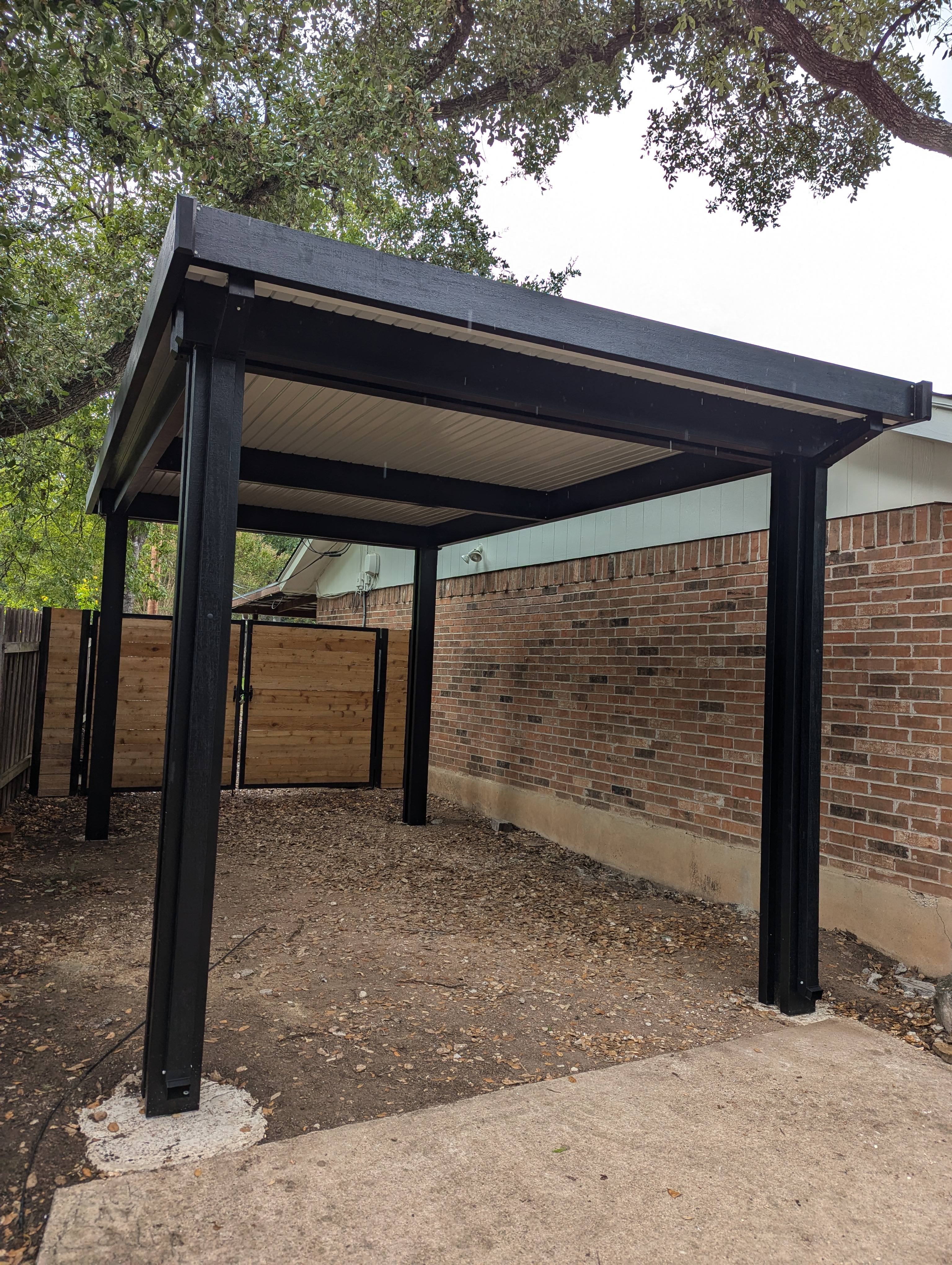 A freestanding carport with a black powder coated steel frame and a pale insulated underside, standing on a concrete and gravel drive alongside a brick house, with a new timber fence and gate behind it.