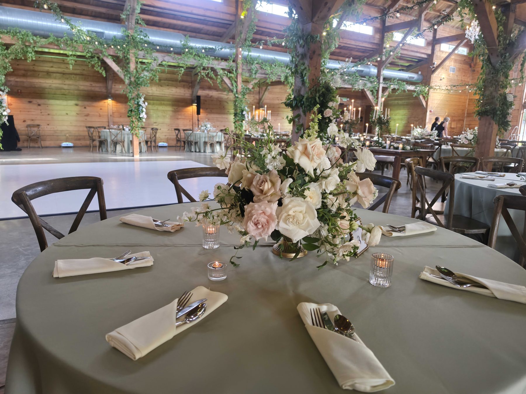 A round table dressed with a floral centerpiece and place settings, in a timber-framed event room with greenery wound around the posts.
