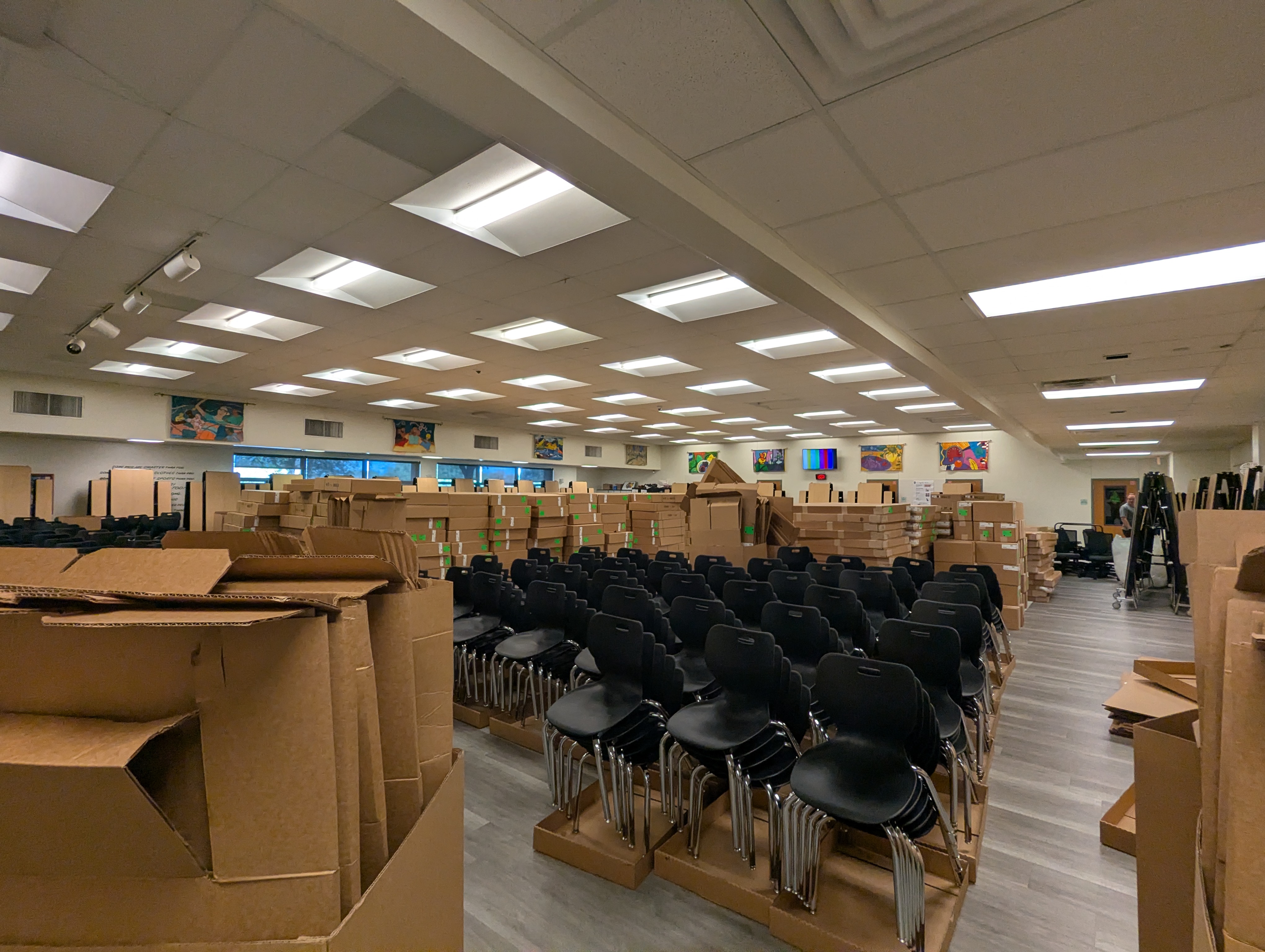 A school library being used as a staging floor, stacked rows of new classroom chairs and hundreds of flat boxes laid out across the room during a campus furniture install.