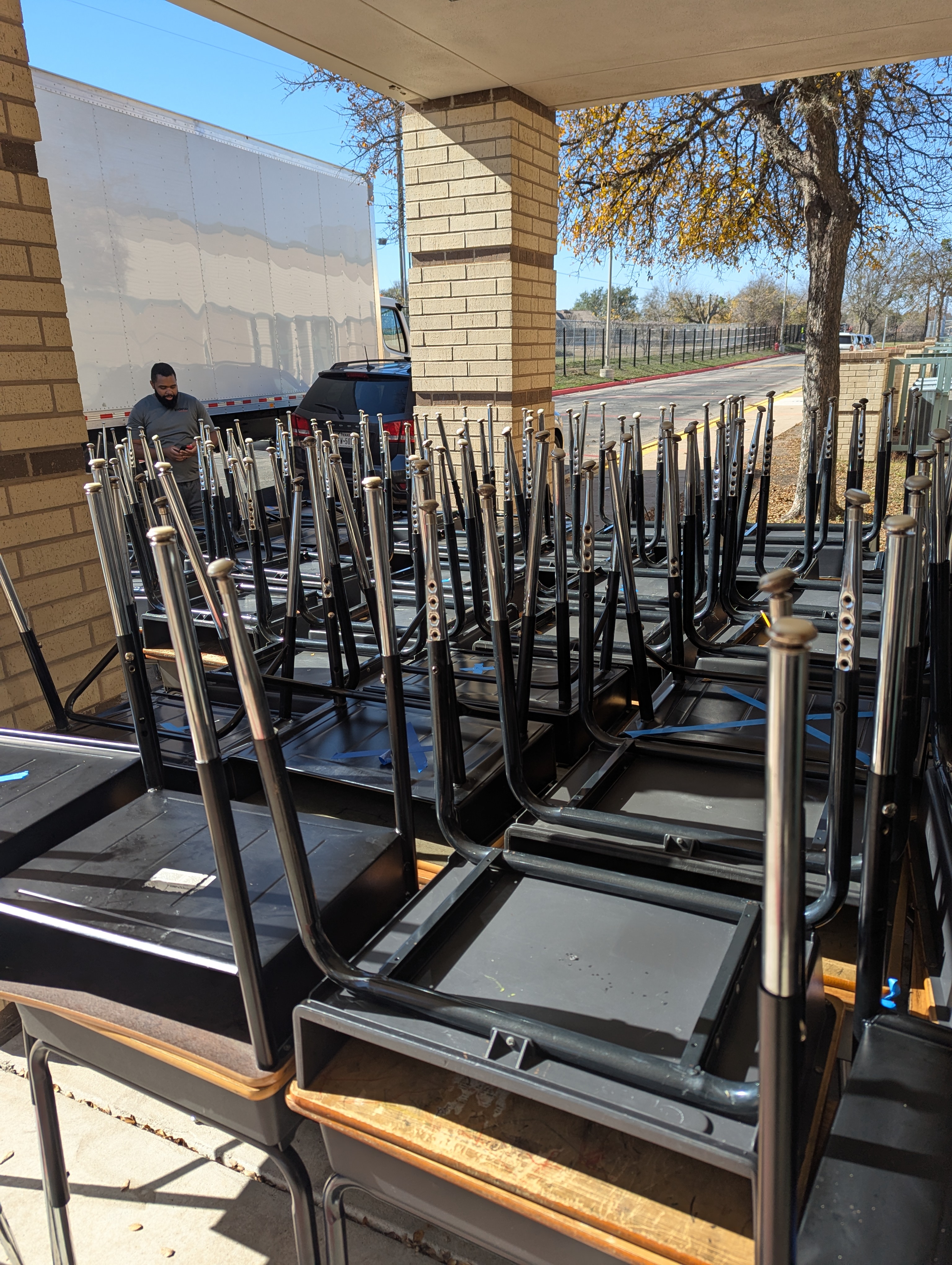 Classroom desks and chairs stacked and staged under a covered walkway beside a box truck, ready to be loaded out.