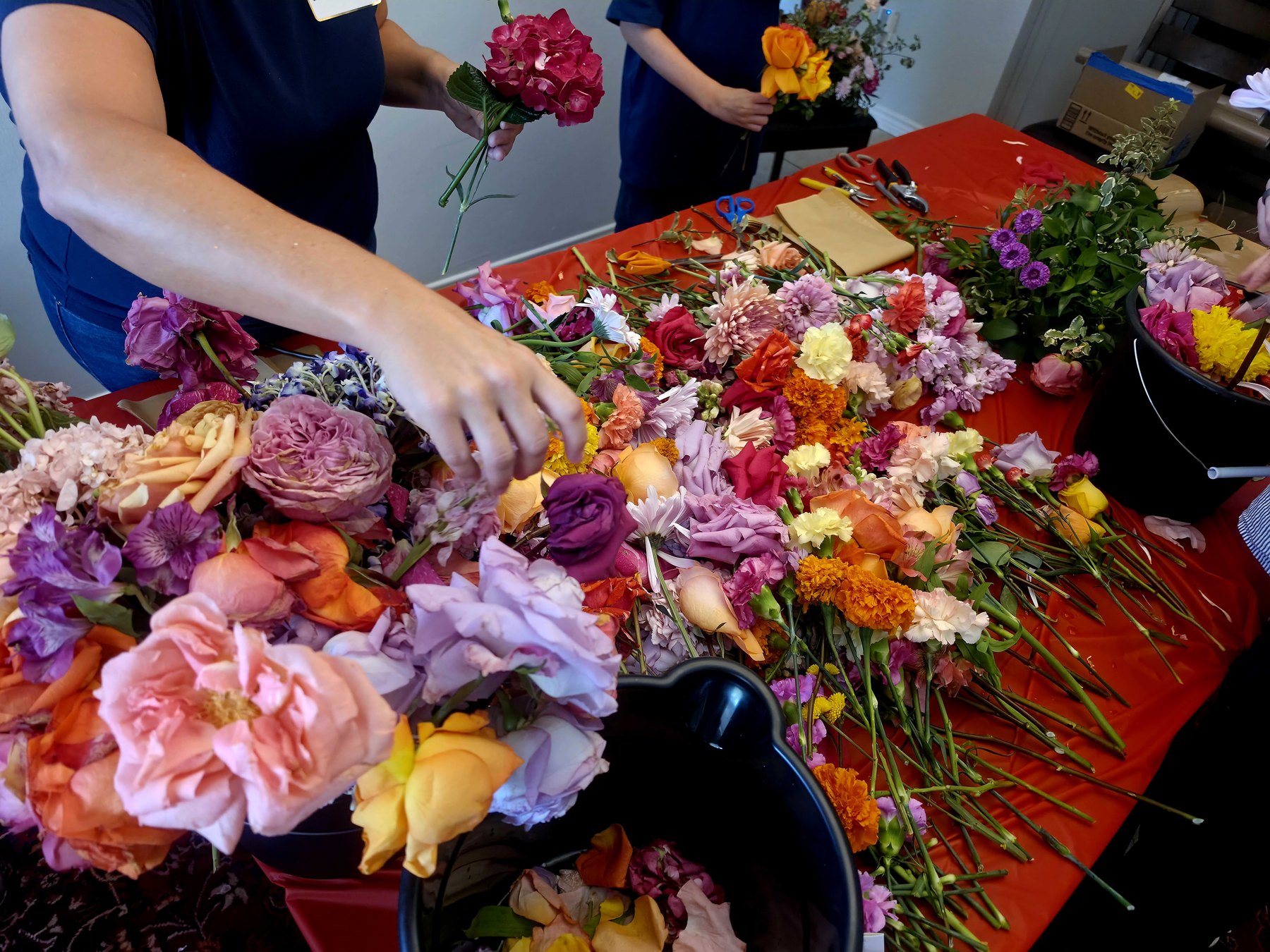 A long table spread with hundreds of cut flowers, roses, carnations, marigolds and hydrangea in pink, orange and cream. Two people are sorting them into small bouquets by hand, with shears, brown paper and black buckets alongside.