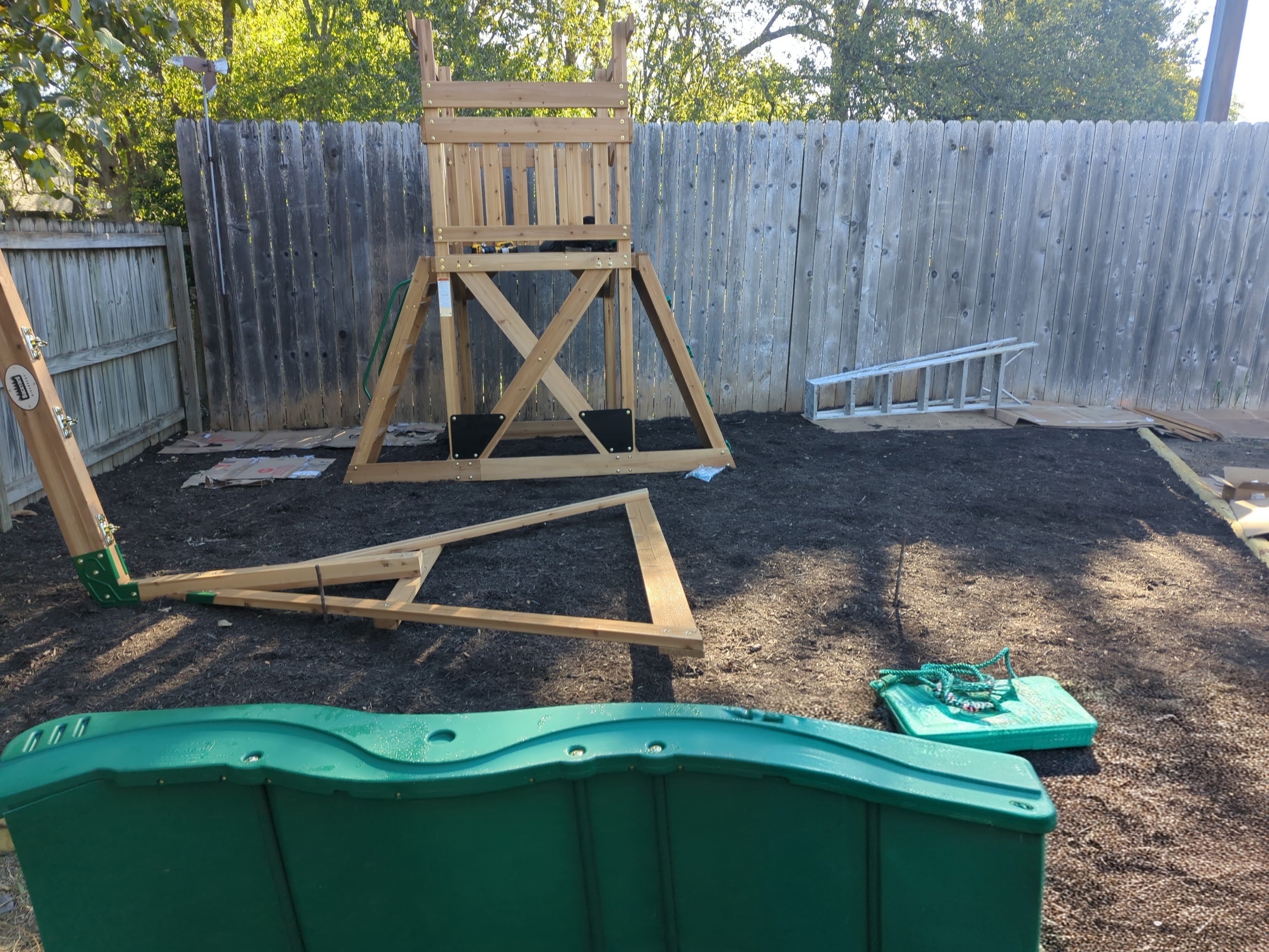 A part built cedar playset tower standing on fresh dark mulch in a fenced backyard, with the swing beam timbers laid out on the ground beside it, a green wave slide waiting in the foreground and flattened packaging stacked against the fence.