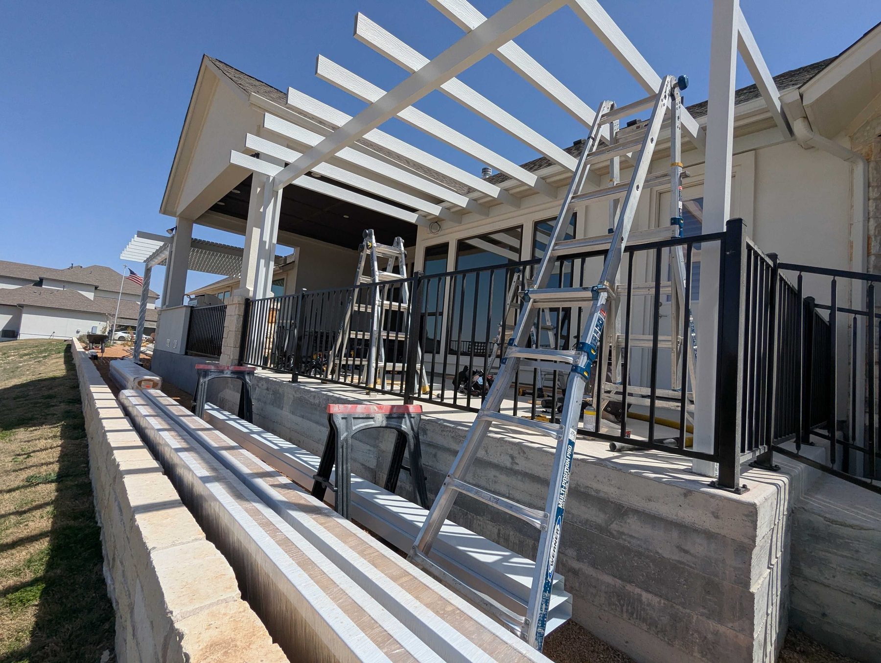 A white pergola frame going up over a raised terrace, rafters in place and a stepladder standing against it.