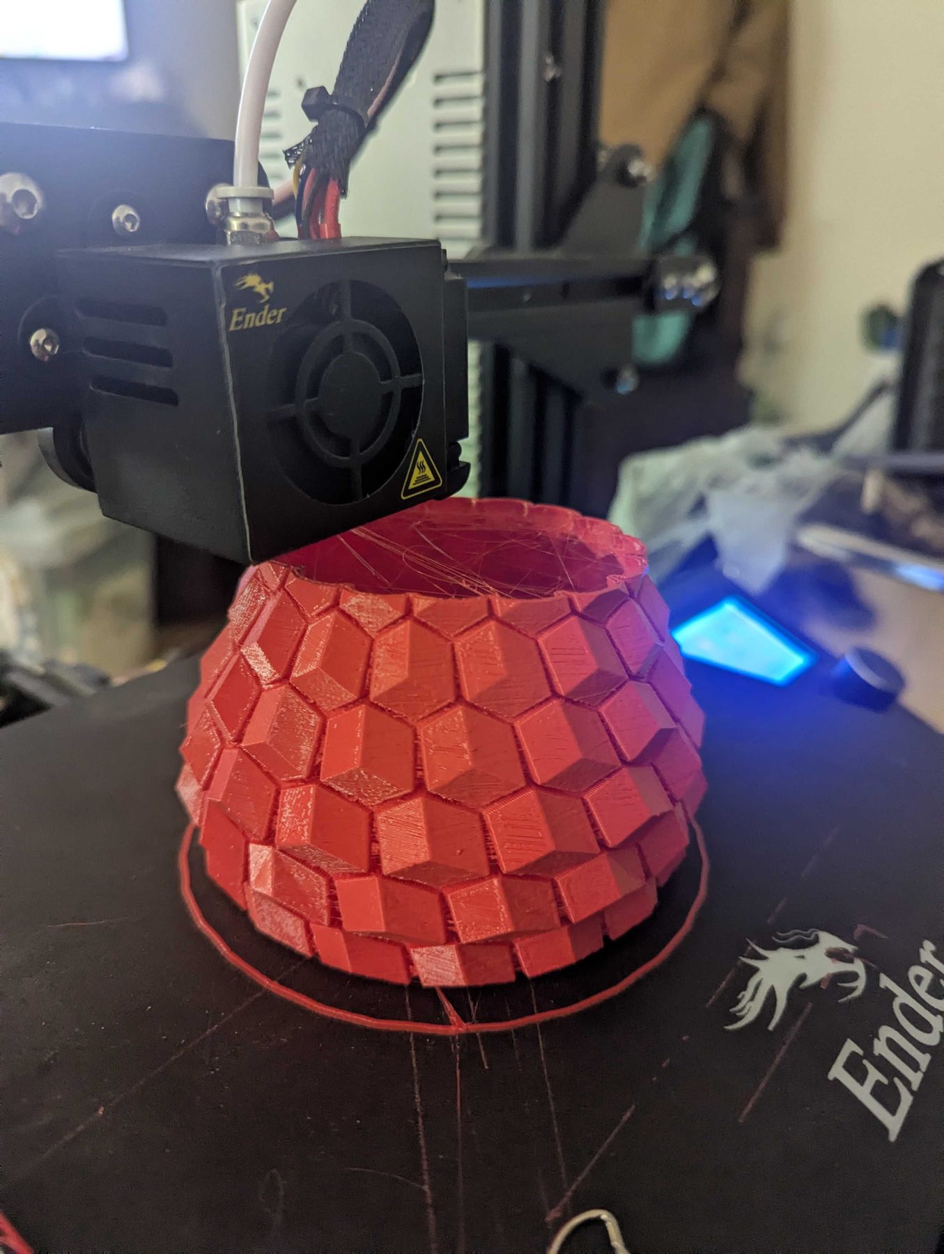 A 3D printer part way through printing a red vessel with a scaled surface, the bed lit blue.