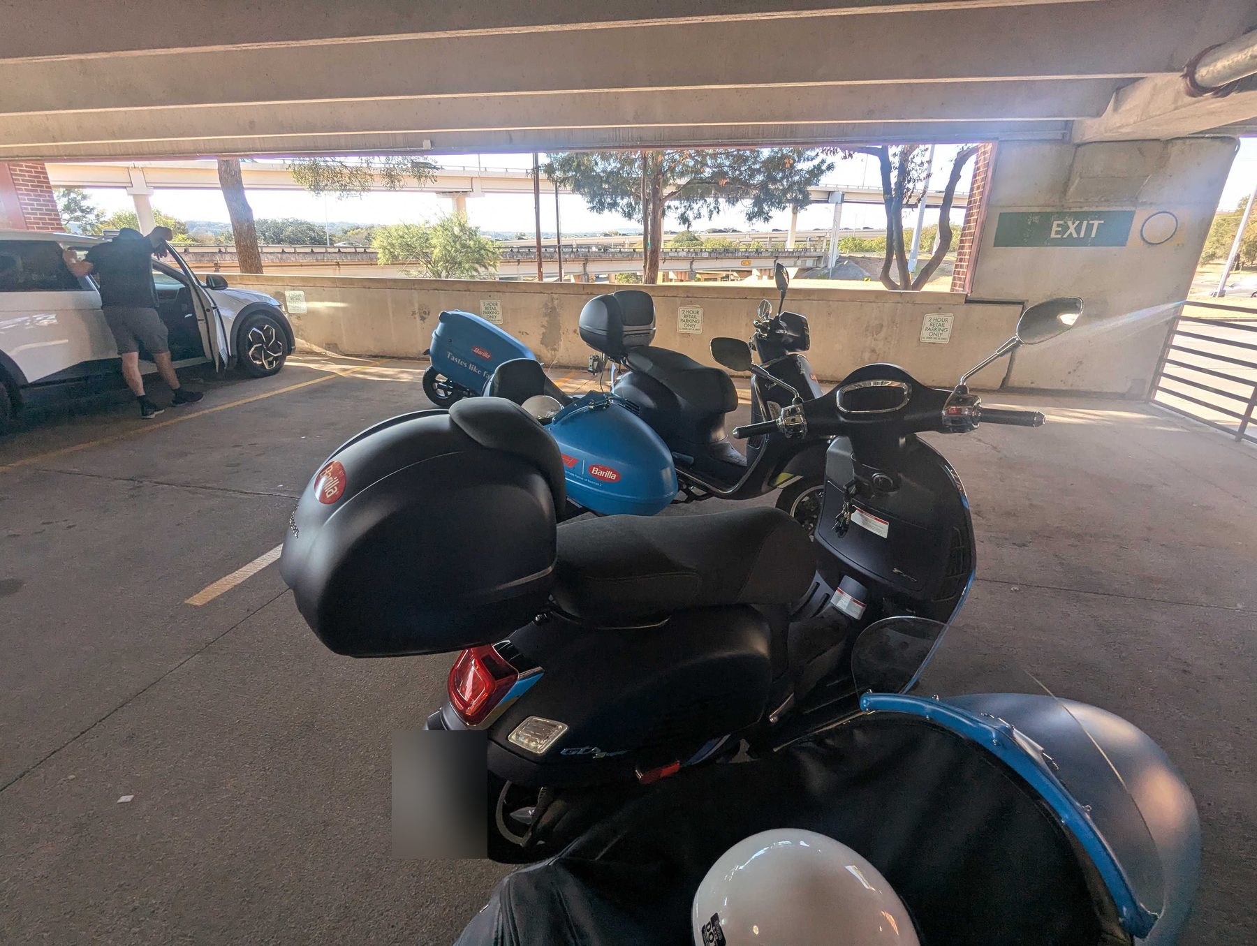 Two scooters parked under a covered deck, each fitted with a hard rear cargo case, a white helmet resting on one seat.
