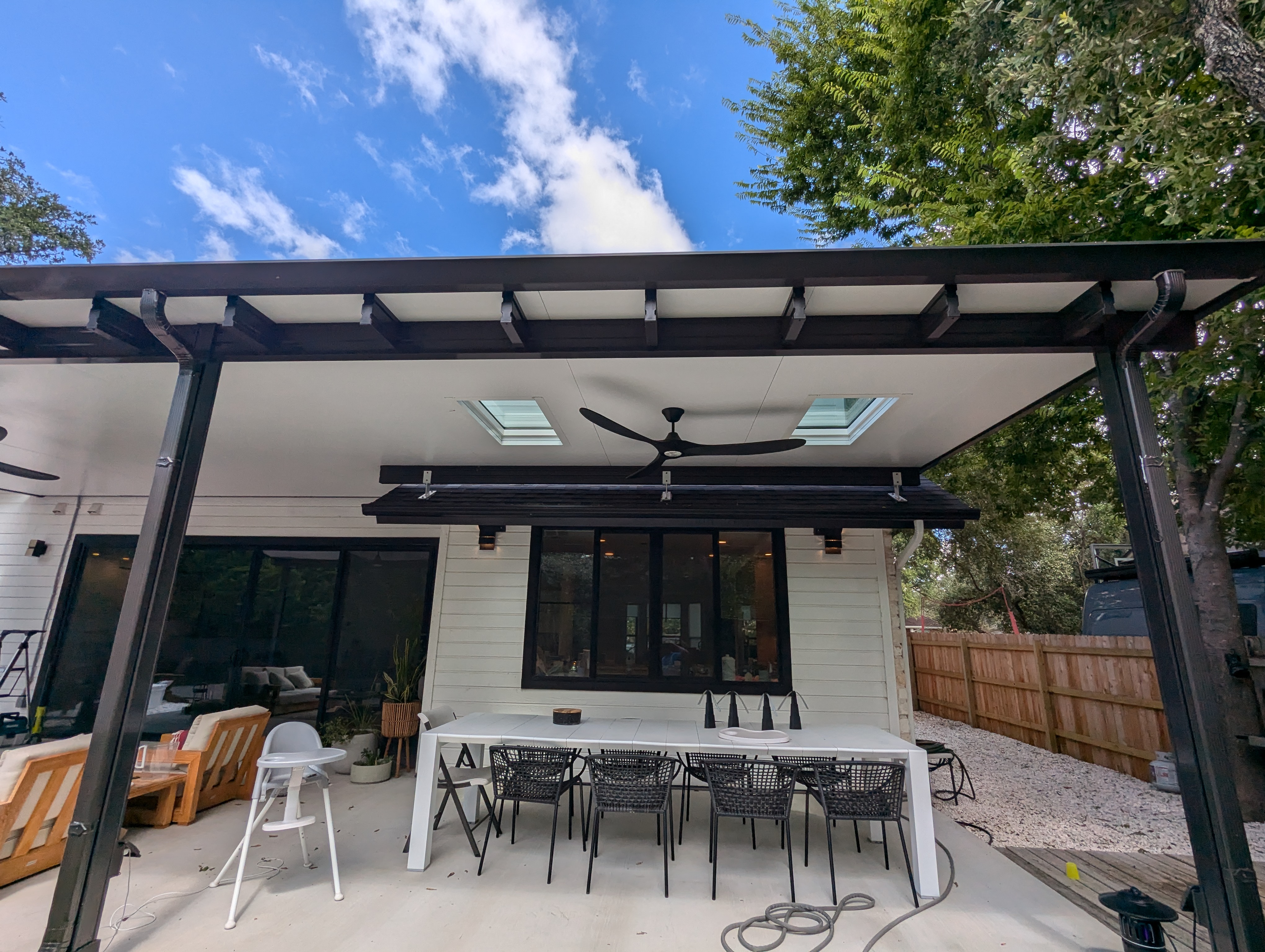 A dark bronze patio cover with an insulated white underside, two skylights and a ceiling fan, attached to a white weatherboard house over a dining table set for eight.