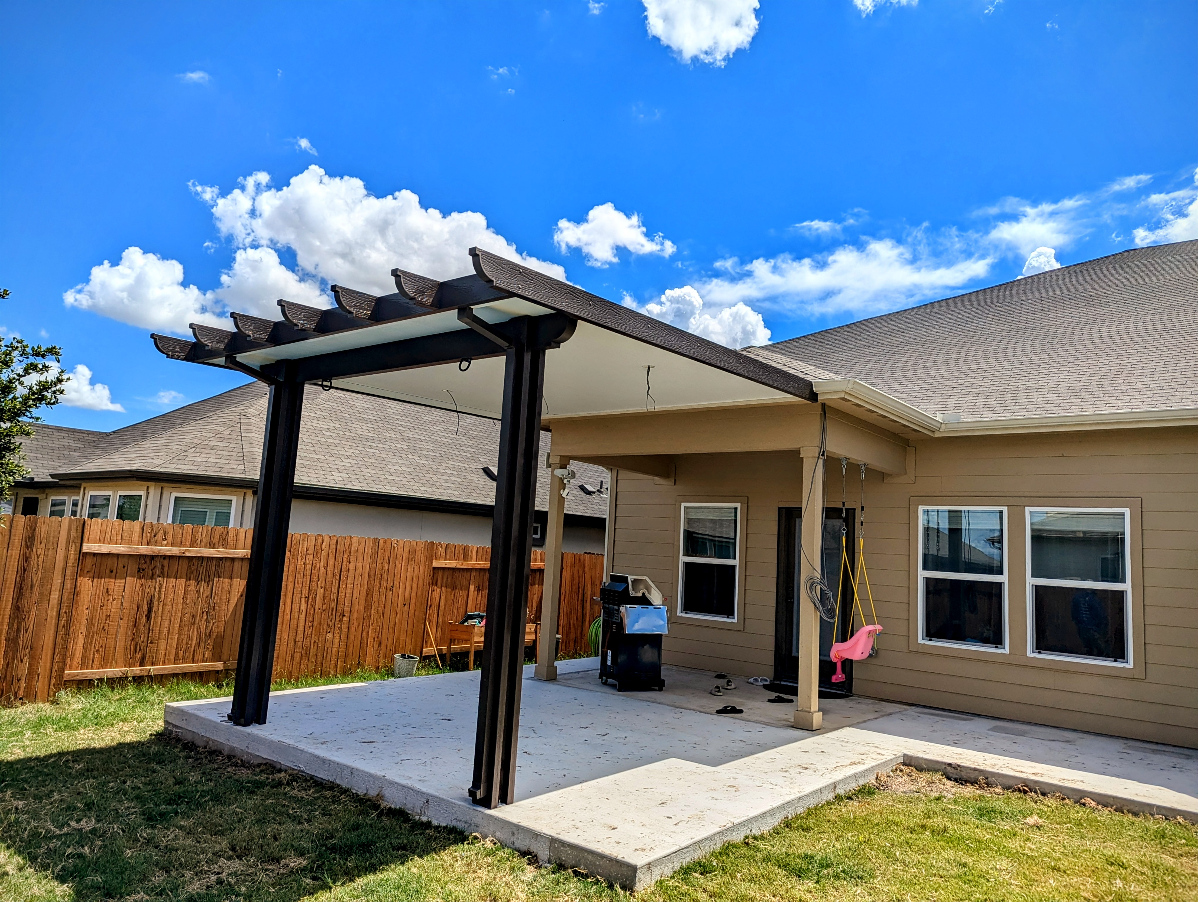 A dark bronze pergola style patio cover with shaped rafter tails along its front edge, attached to a tan house and shading a concrete patio, under a bright blue sky with white cloud.