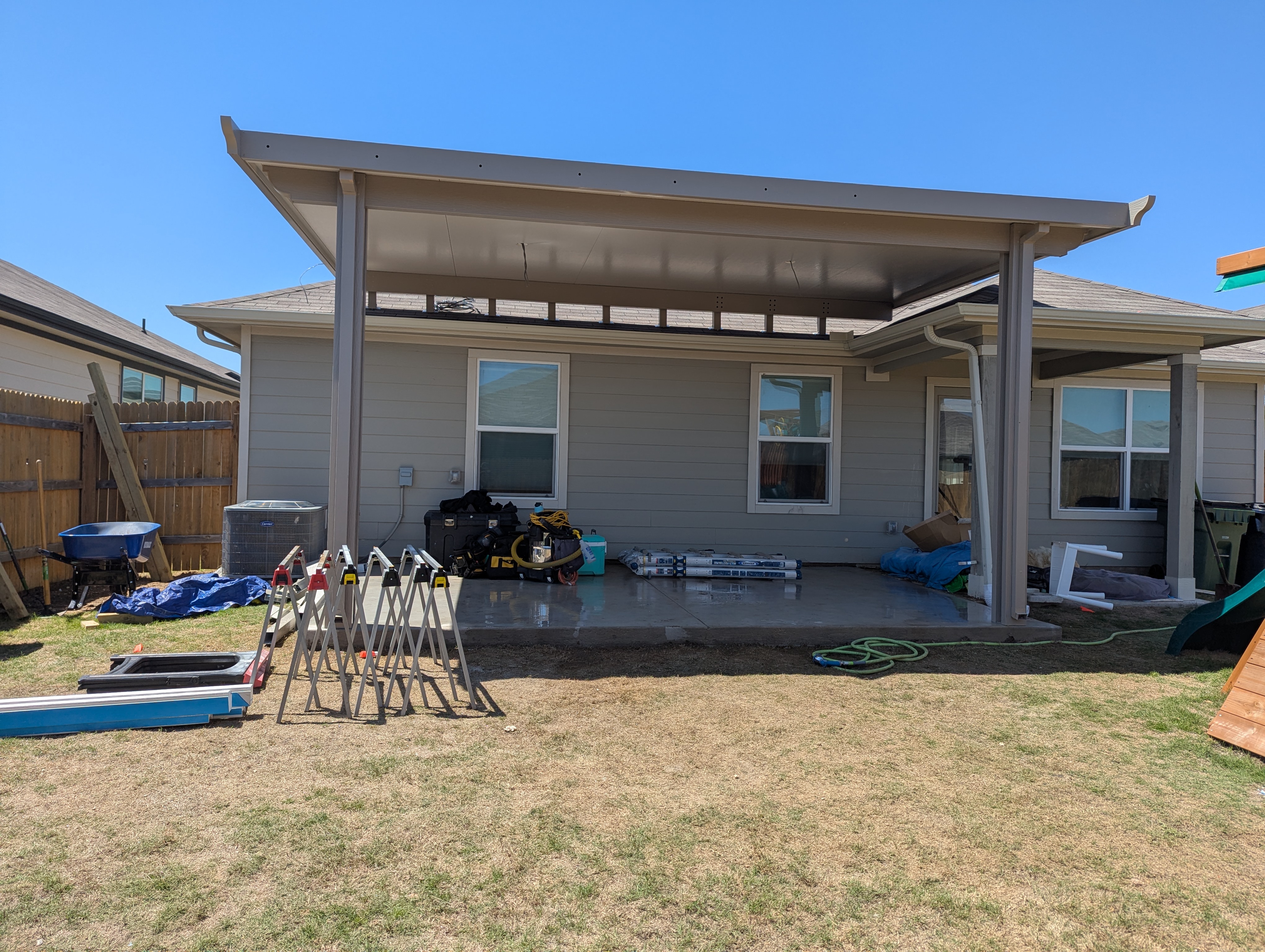 A wide beige insulated patio cover attached above the roofline of a single story house, shading a newly poured concrete slab, with sawhorses and materials still set out on the grass.