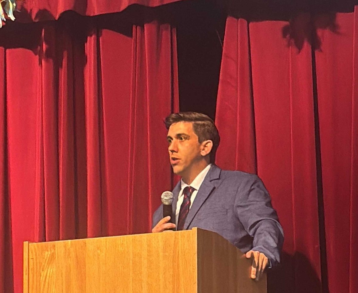 Brendon Wash speaking at a wooden lectern with a handheld microphone, mid-sentence, in a blue suit and tie against a red stage curtain.