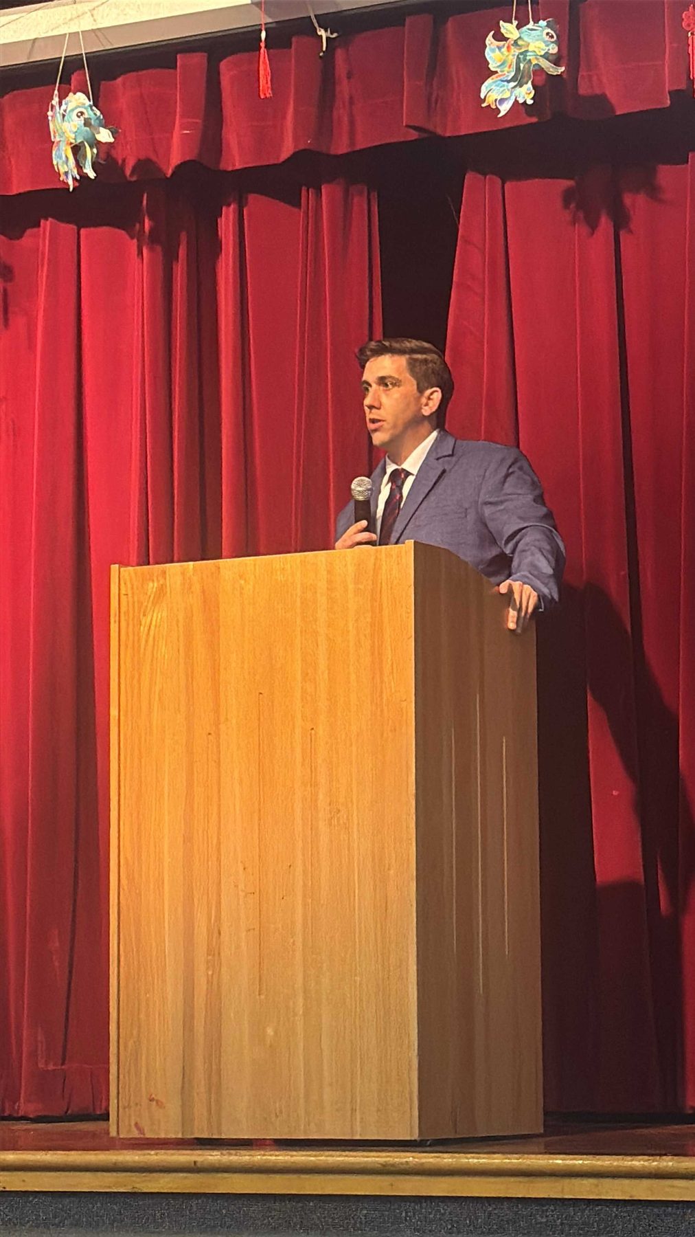 A speaker standing at a wooden lectern on a stage in front of a red curtain.