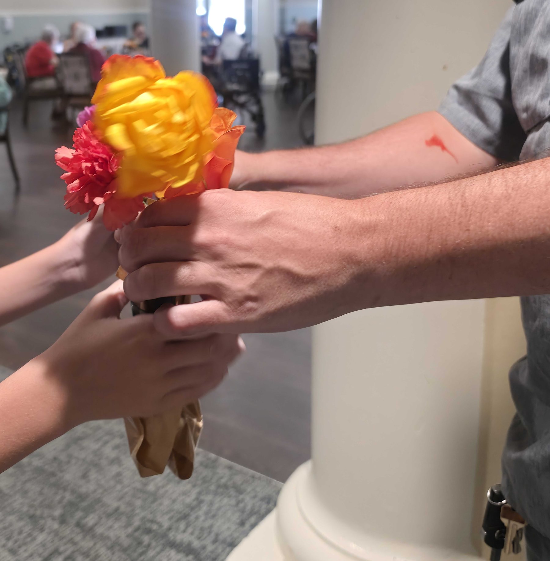 A child's hands and an adult's hands together around a posy of yellow and pink flowers wrapped in brown paper.