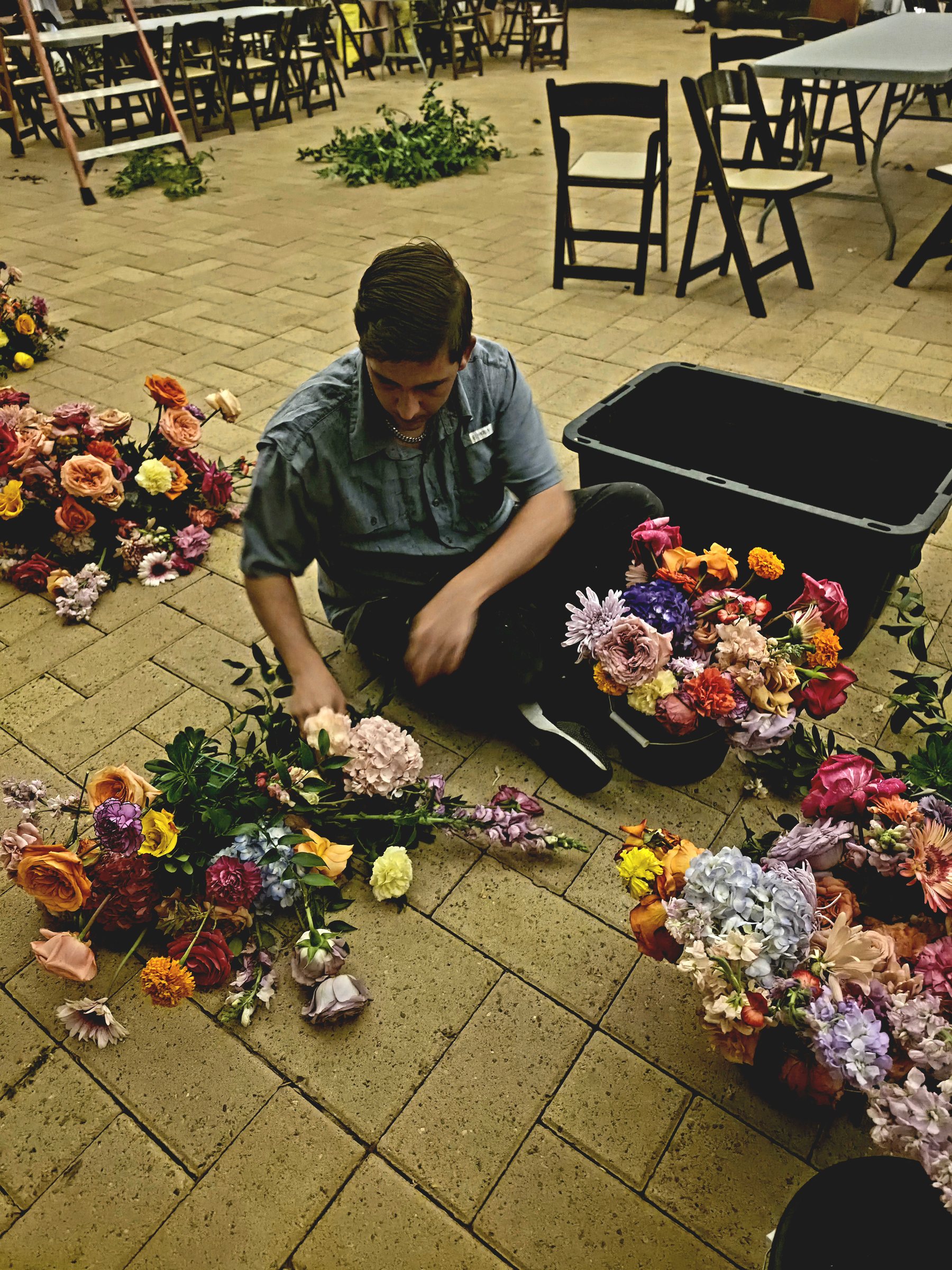 Flowers laid out in piles on a venue floor after a wedding breakdown, being sorted into a bin beside stacked folding chairs.