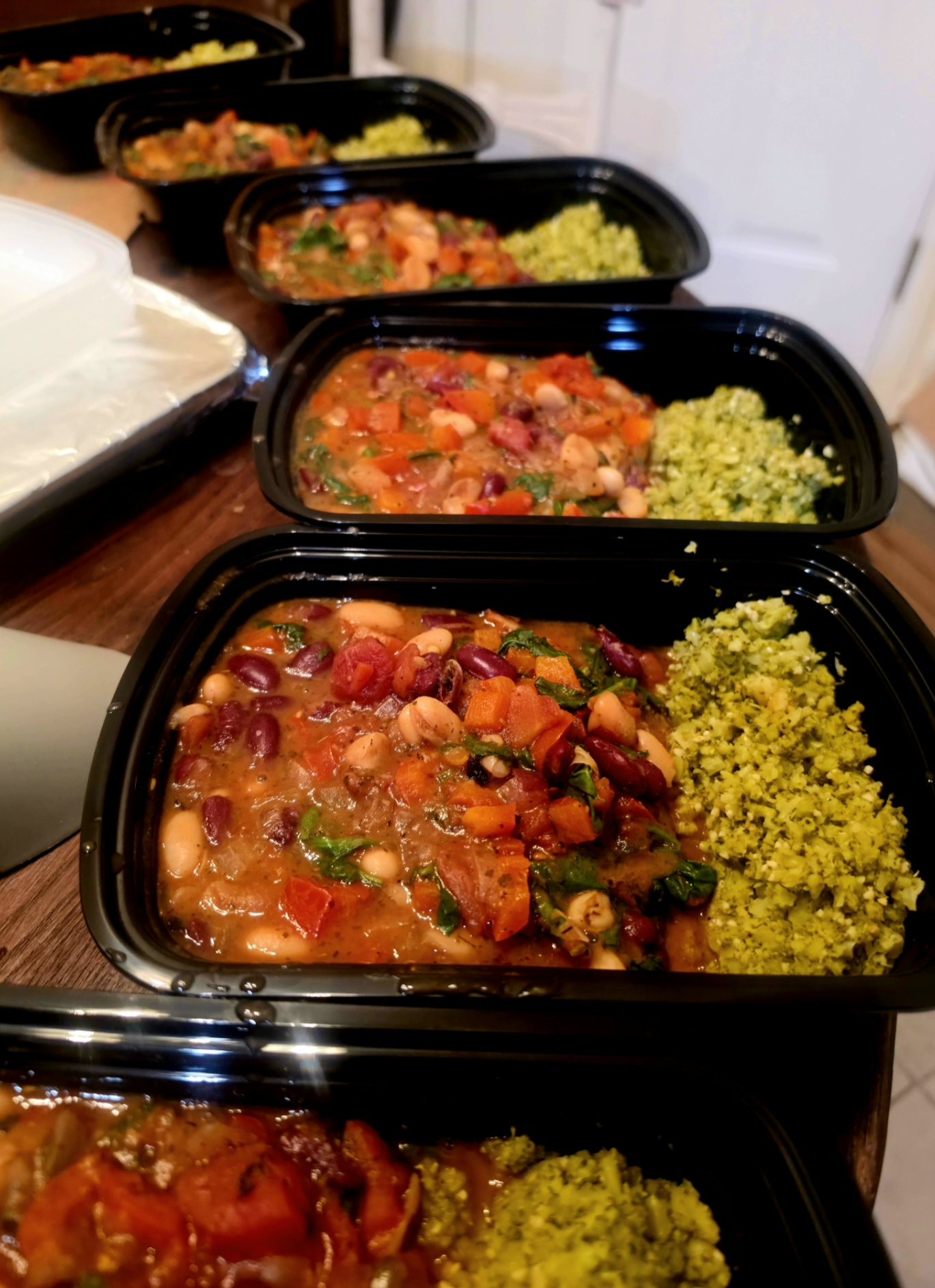 Six meal containers open in a row, each holding a bean stew of kidney beans, white beans and black-eyed peas with carrot, red pepper and spinach, and a portion of herbed broccoli rice.