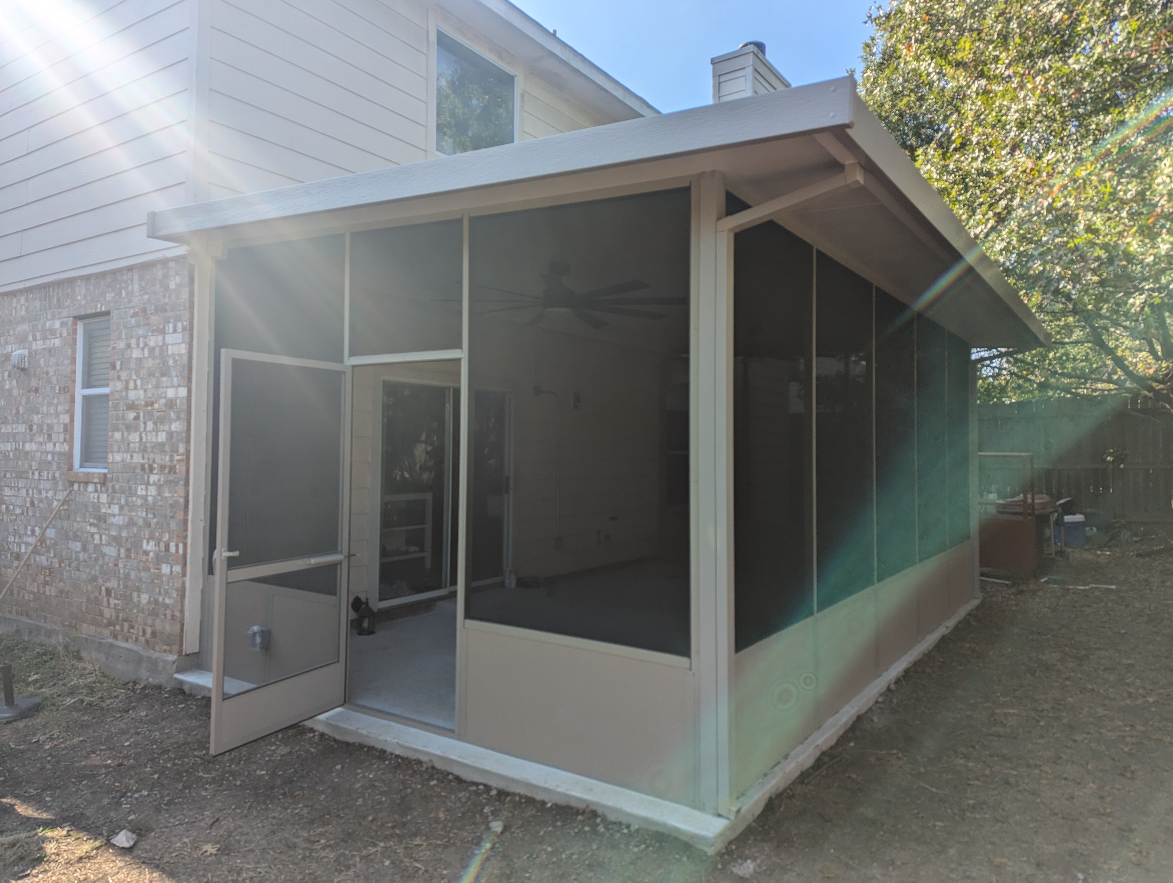 A finished screen room built onto a brick house, with a sloped beige roof, full height screen panels, a screen door standing open and a ceiling fan visible inside.