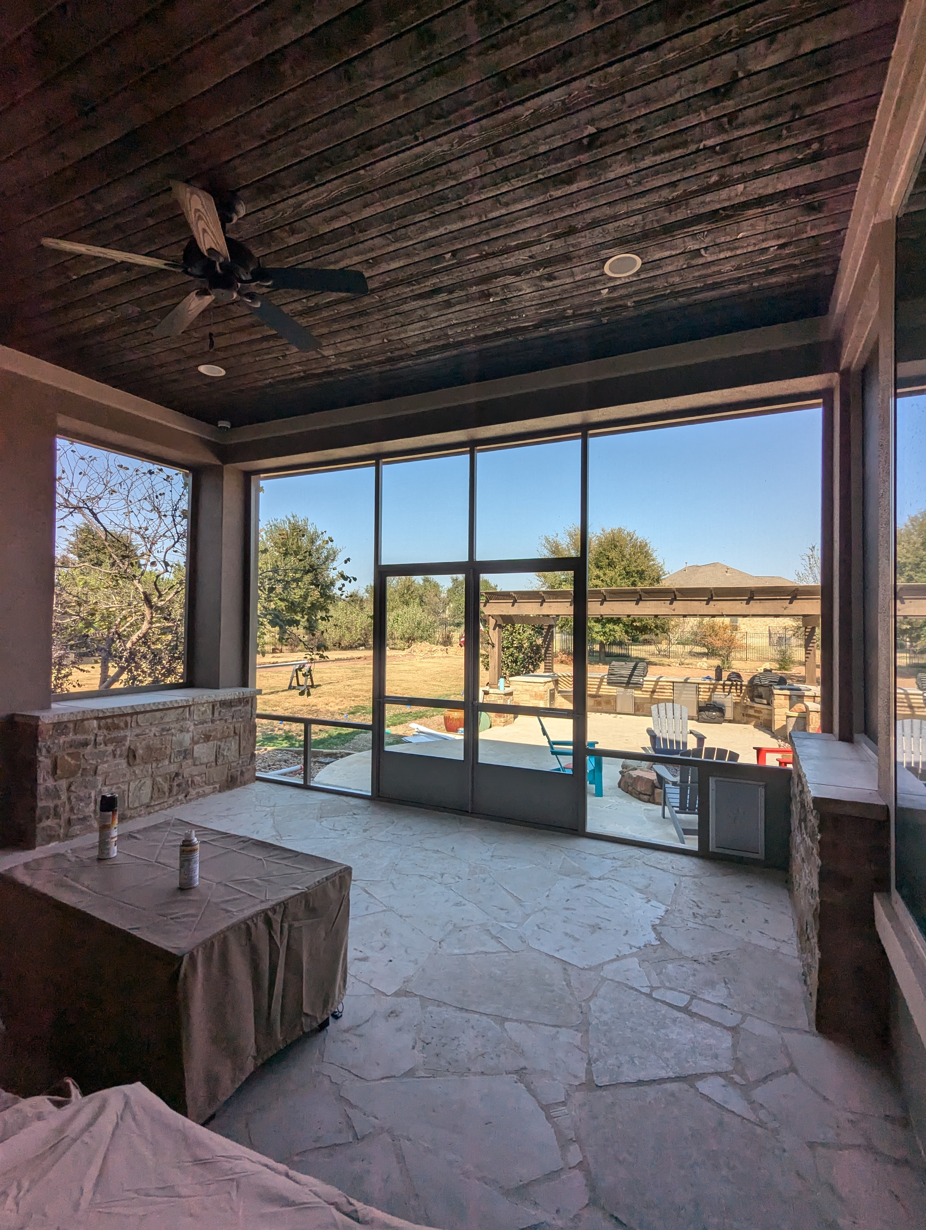 The inside of a screen room looking out through full height screen walls to a pool terrace, with a stained timber ceiling, a ceiling fan and a stone knee wall.