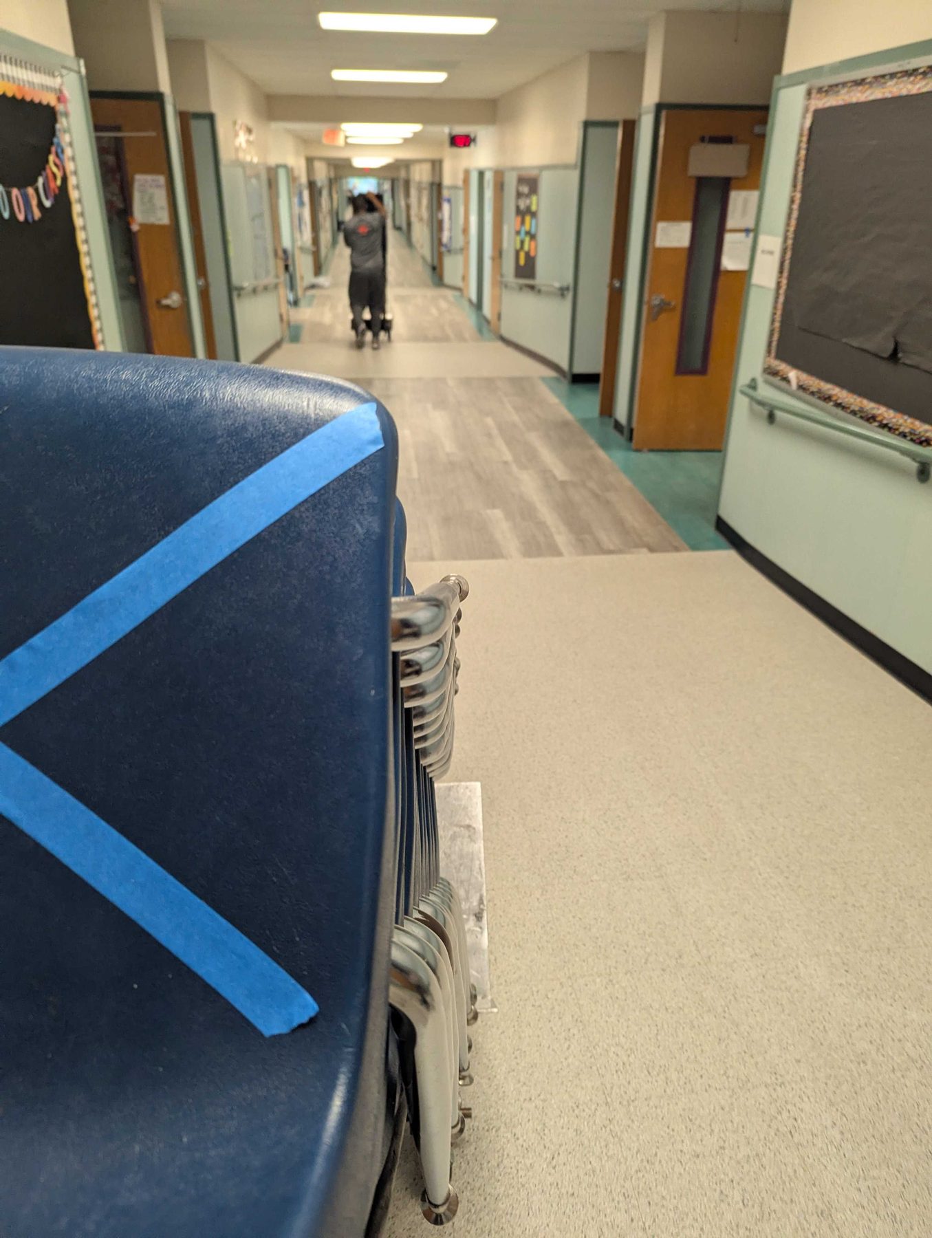 A school corridor with classroom doors down both sides and a trolley of stacked chairs pushed into the foreground.